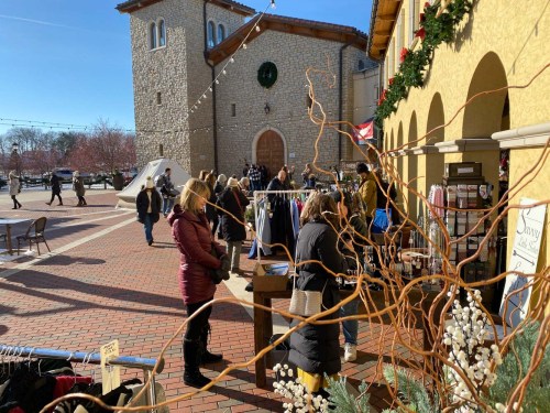 A bustling outdoor market scene with shoppers browsing clothing stalls near a festive building decorated for the holidays.