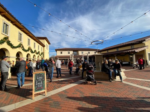 A bustling outdoor market scene with people mingling, string lights overhead, and a fountain in the center.