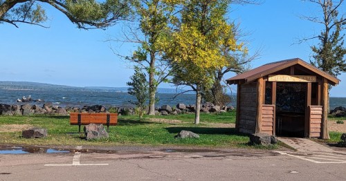 A wooden shelter near a lake, with a bench and trees, under a clear blue sky. Waves can be seen in the background.