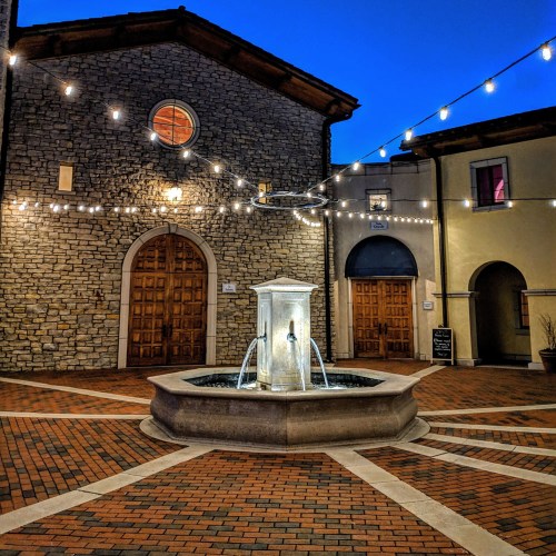 A charming courtyard with a fountain, surrounded by stone buildings and illuminated by string lights at dusk.