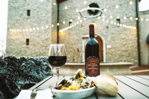A glass of red wine and a plate of food on a table, with a stone building and string lights in the background.
