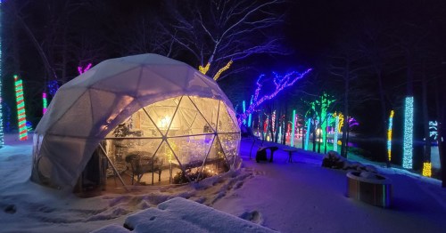 A glowing geodesic dome surrounded by colorful lights in a snowy landscape at night.