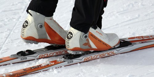 Close-up of ski boots on skis, set against a snowy background. The boots are white and orange, labeled "Rossignol."