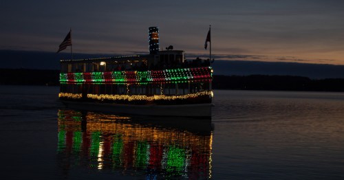 A brightly lit boat with colorful lights glides across a calm lake at dusk, reflecting on the water.