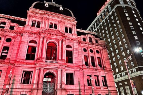 Historic building illuminated in red at night, with a tall hotel visible on the right.