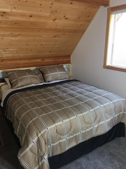 Cozy bedroom with a wooden ceiling, featuring a double bed with a patterned quilt and natural light from a window.