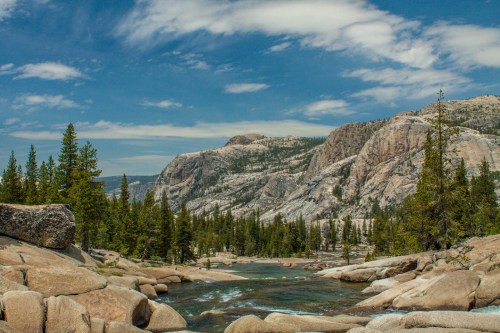 A scenic view of a rocky landscape with a river, surrounded by tall trees and mountains under a blue sky with clouds.