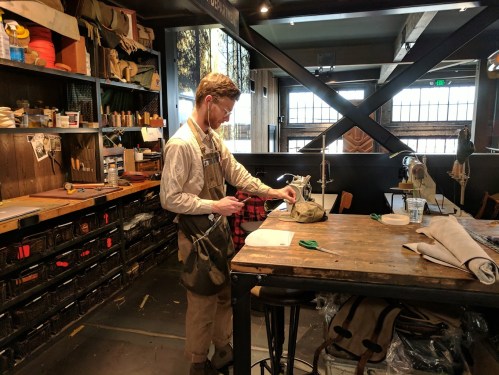 A craftsman works at a wooden table in a workshop, surrounded by sewing machines and tools.