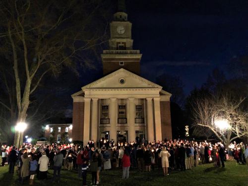 A large group of people holding candles gathers in front of a historic building at night.