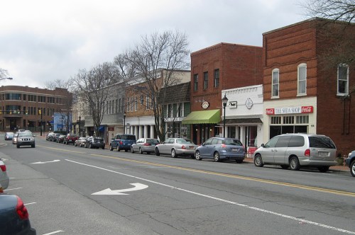 A street lined with brick buildings and shops, featuring parked cars and a cloudy sky overhead.