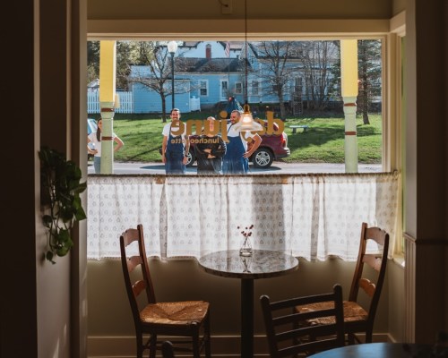 View from inside a café, showing two people outside and a table set for two by the window.