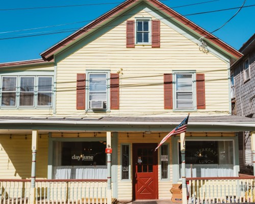 A yellow two-story building with red shutters, featuring a sign for breakfast and lunch, and an American flag out front.