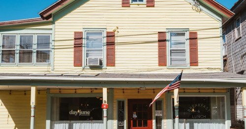 A yellow two-story building with red shutters, featuring a sign for breakfast and lunch, and an American flag.