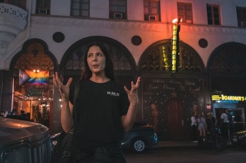 A woman gestures expressively in front of a brightly lit building at night, with colorful signs and decorations.
