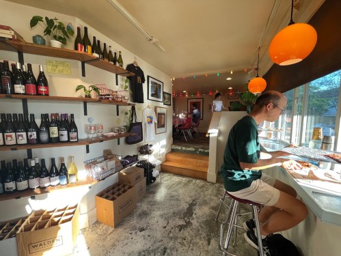 A cozy wine bar with shelves of bottles, a person sitting at a counter, and a dining area with festive lighting.