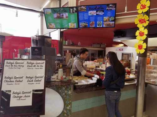 A customer orders food at a counter with a menu display and specials sign in a vibrant food market setting.