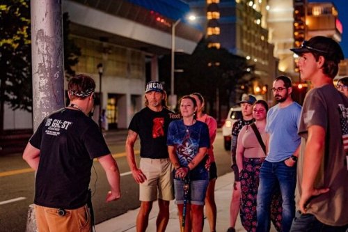 A group of people gathered on a city street, listening to a speaker near a lamppost at dusk.