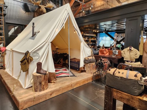 A cozy camping tent display with rustic decor, surrounded by outdoor gear and clothing in a stylish indoor setting.