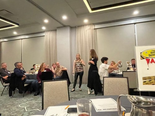 A group of people at a dinner event, with two women standing and engaging in a presentation or performance.
