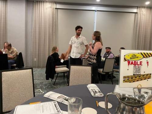 A man and woman converse at a round table in a conference room, with other attendees in the background.
