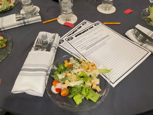 A salad with lettuce, tomatoes, croutons, and dressing on a table set with utensils and a menu.
