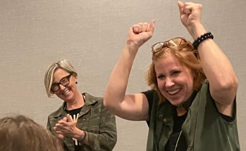 Two women celebrate joyfully, one clapping and the other raising her arms in excitement, against a neutral background.