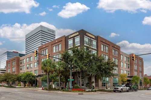 A modern brick hotel with palm trees, located at an intersection, under a blue sky with clouds.