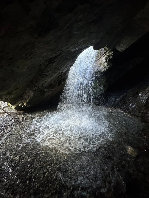 A waterfall cascades from a rocky overhang into a pool below, surrounded by dark stone and pebbles.