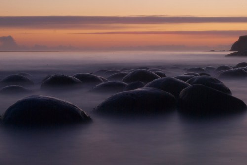 Smooth, rounded stones partially submerged in calm water at sunset, with a soft, colorful sky in the background.