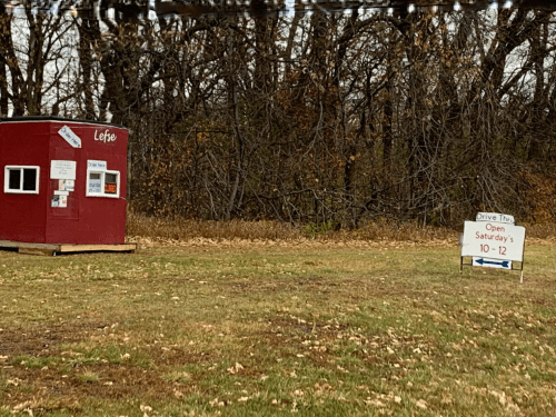 A small red shed with signs, surrounded by trees, and a sign indicating it's open Saturdays from 10 to 12.