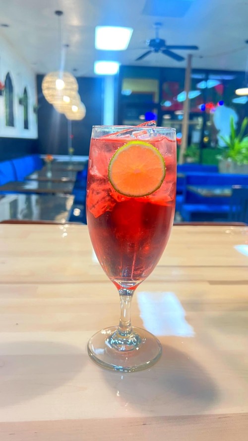 A tall glass filled with a red drink, ice, and a slice of lime, set on a wooden table in a cozy restaurant.