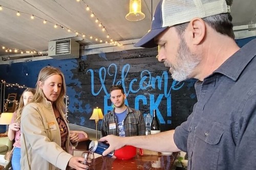 A man pours a drink for a woman at a bar, while another man watches. The setting has a welcoming atmosphere with lights.