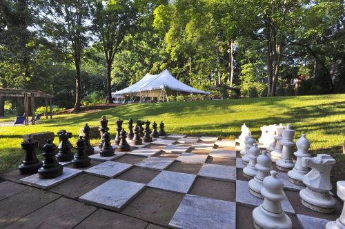 A large outdoor chess set with black and white pieces on a stone board, set in a green garden with a tent in the background.