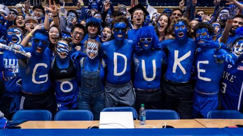 A lively crowd of fans with blue body paint, cheering and holding up letters spelling "GO DUKE" at a sports event.