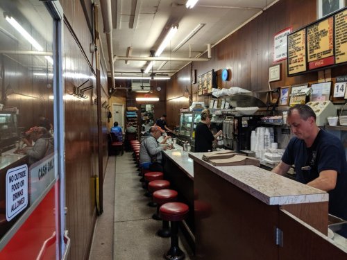 A diner interior with a long counter, red stools, and patrons seated, while a staff member prepares food behind the counter.