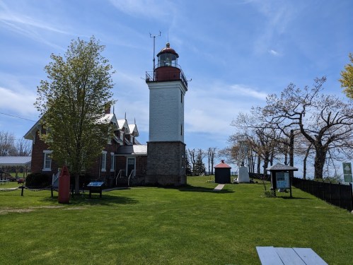 A historic lighthouse beside a brick building, surrounded by green grass and trees under a clear blue sky.