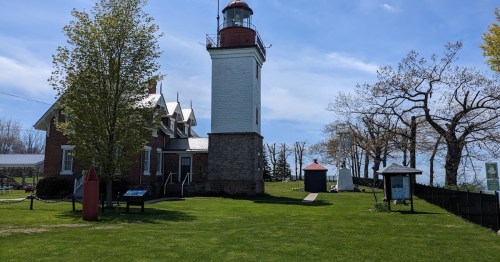 A historic lighthouse stands beside a grassy area, with trees and a blue sky in the background.
