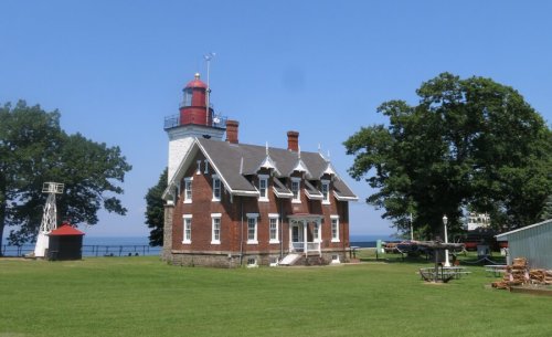 A historic brick lighthouse with a red top, surrounded by green grass and trees, near a body of water under a clear blue sky.