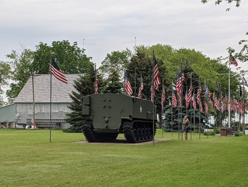 A military vehicle displayed on a grassy area surrounded by American flags and trees, with a building in the background.