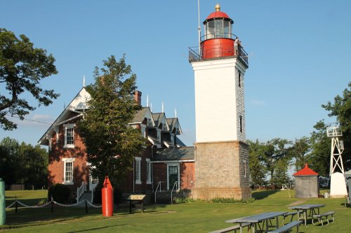 A historic lighthouse with a red top, surrounded by green grass and trees, next to a brick building and picnic tables.