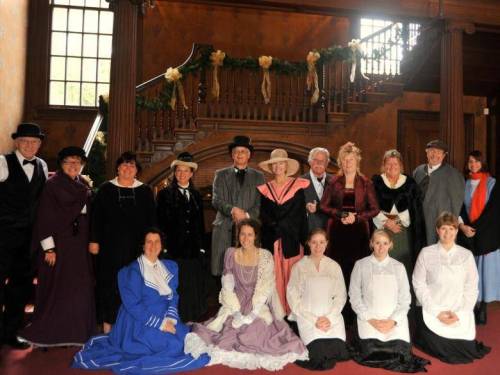 A group of people in historical costumes poses together in a grand interior with festive decorations.