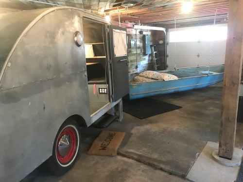 A vintage camper and a small blue boat inside a garage, with a welcome mat on the floor.