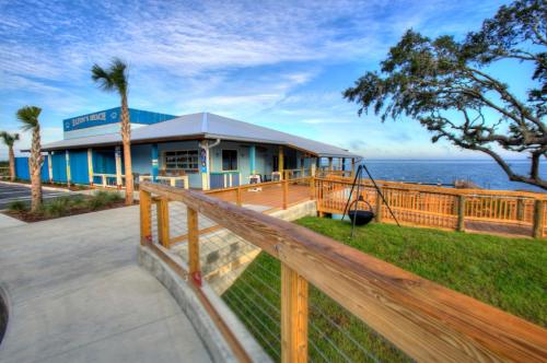 A coastal building with a wooden deck, palm trees, and a swing, overlooking the water under a blue sky.