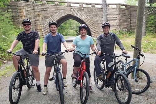 Four people on bicycles pose in front of a stone bridge in a wooded area, smiling and wearing helmets.