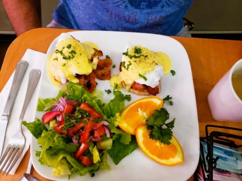 A plate of eggs Benedict with bacon, hollandaise sauce, and a side salad of lettuce, tomatoes, and orange slices.