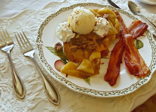 A plate of warm apple dessert topped with ice cream and whipped cream, alongside crispy bacon strips.