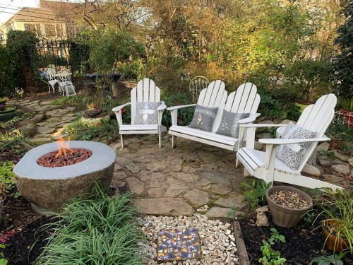 A cozy garden seating area with white Adirondack chairs, decorative pillows, and a stone fire pit surrounded by greenery.