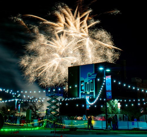 Fireworks illuminate the night sky over Enid, Oklahoma, with festive lights and a crowd below.