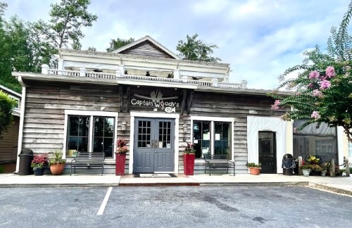 A rustic wooden building with a sign reading "Captain Woody's," surrounded by potted plants and a parking area.