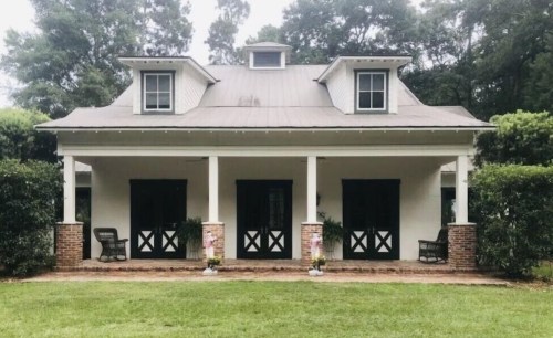 A charming white house with a porch, featuring black doors and rocking chairs, surrounded by greenery.